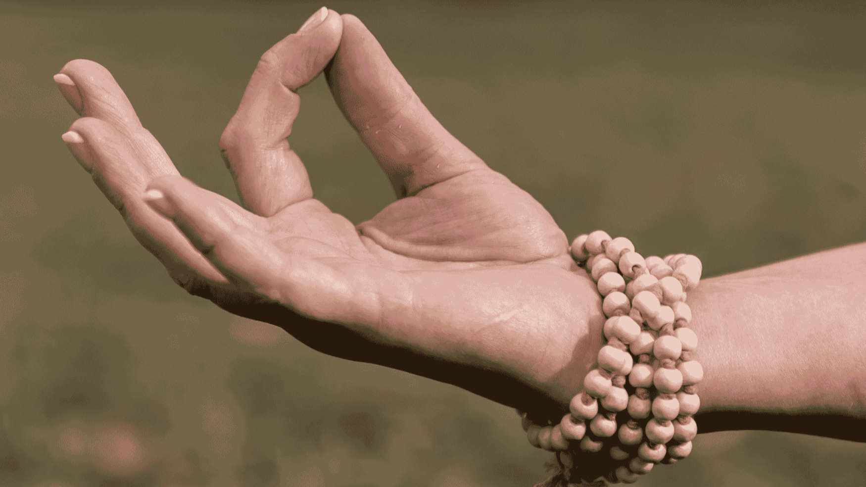Close-up of a person's hand making a mudra gesture, with a beaded bracelet on the wrist, against a blurred background.