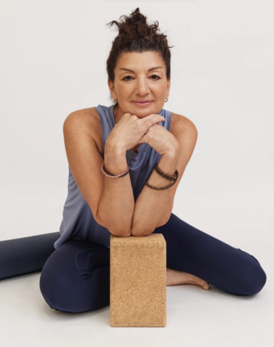 A woman with dark hair tied up in a bun, wearing a blue single top and black yoga pants with her elbows on a cork yoga block sitting on the floor white background looking at the camera