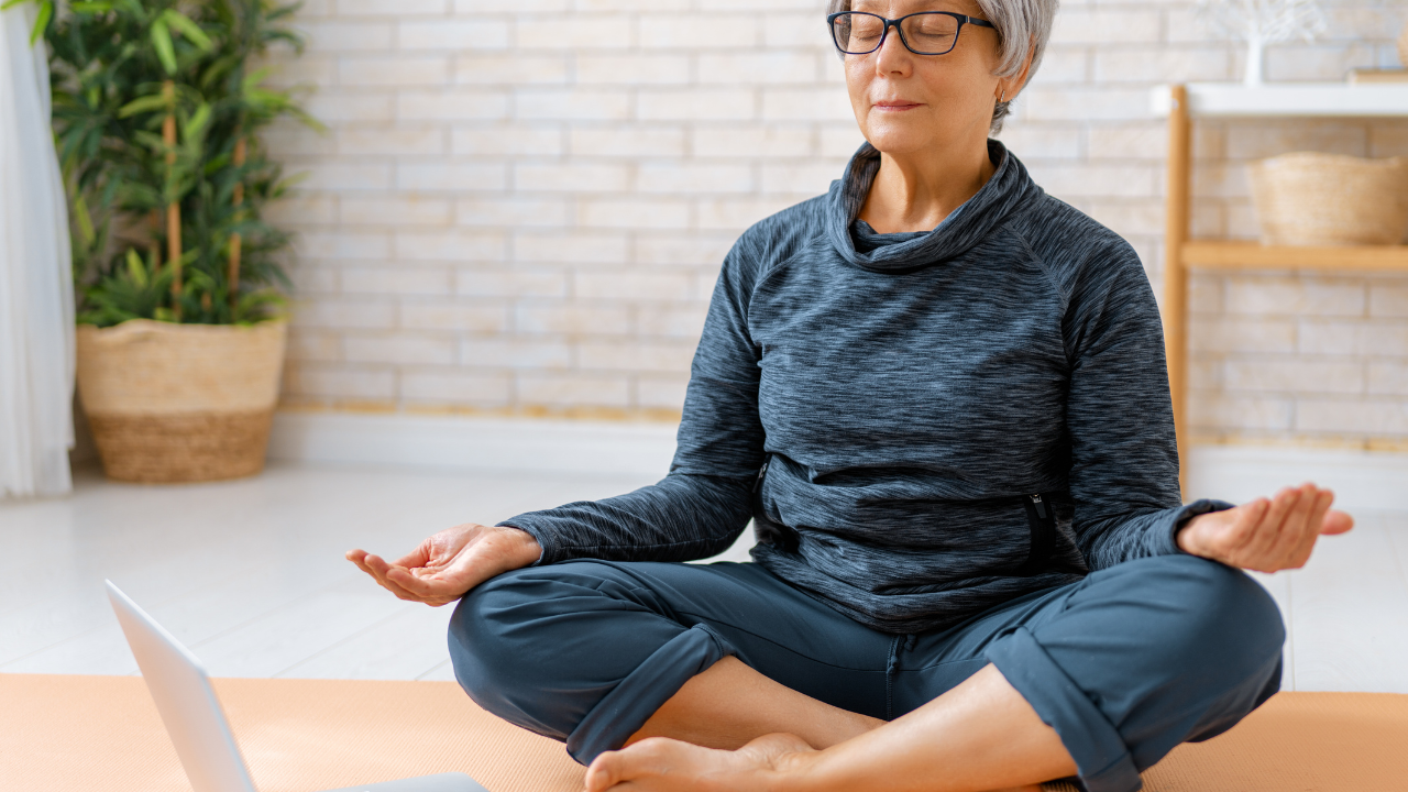 An elderly woman in a gray athletic jacket practicing meditation in a yoga pose on a mat in a bright living room.