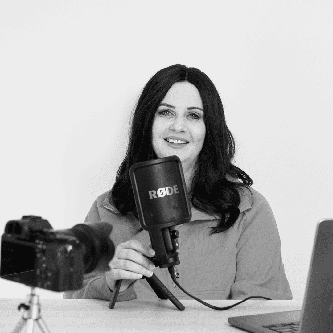 Woman with black hair wearing a jacket, holding a Rode microphone, sitting at a wooden table with a laptop and camera in a room with a plain white wall.