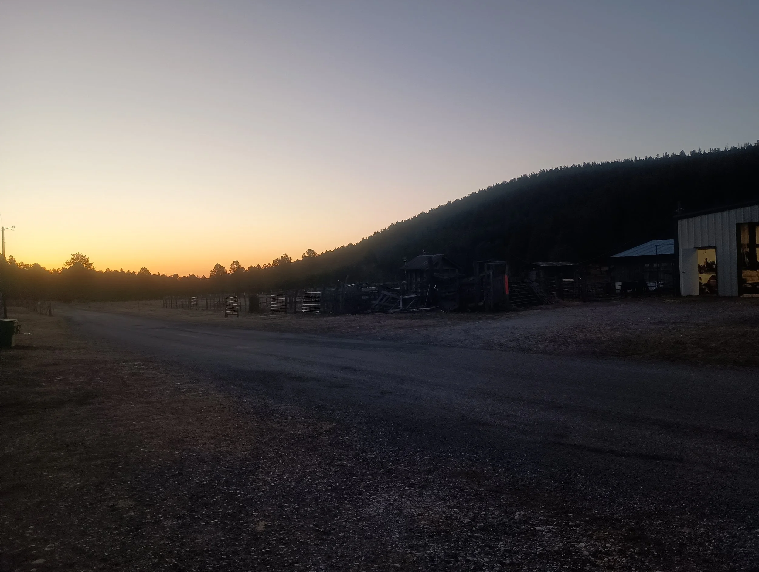 A rural scene at dusk with a dirt road, fencing, a barn, and a hillside covered with trees in the background.