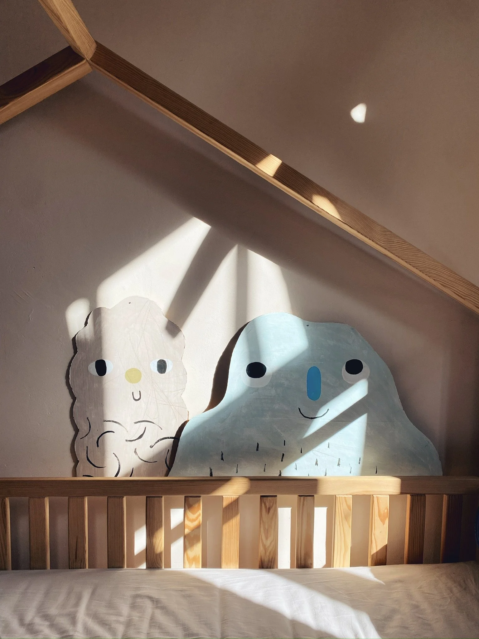 Bett mit einem Holzrahmen und zwei Wandbildern, eines in Form eines blauen Gesichts und das andere wie ein multipliziertes Eis, mit Sonnenlicht und Schatten auf der weißen Wand.