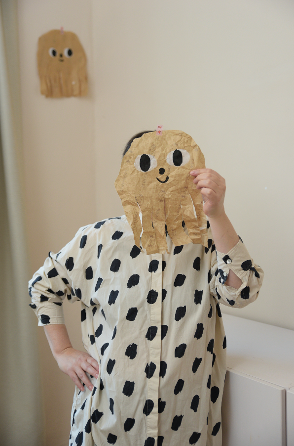 Ruth Biniwersi hält ein Papercut Monsterchen mit einem lachenden Gesicht vor das Gesicht, während im Hintergrund eine weitere Papercut Figur mit einem lachenden Gesicht an der Wand hängt.