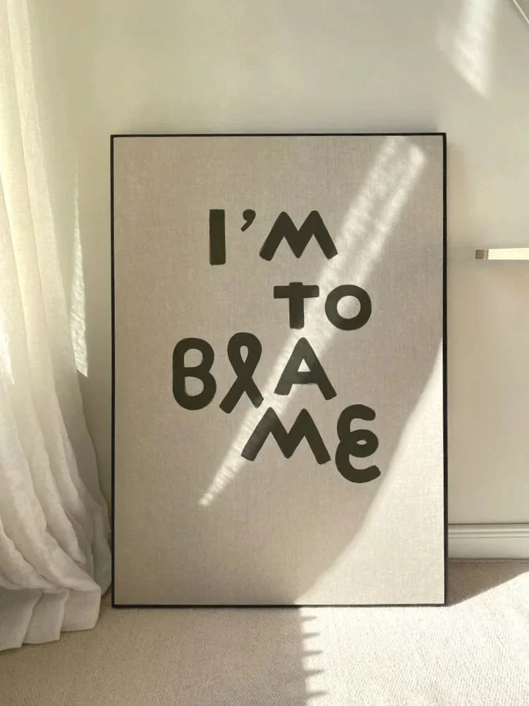 Ein Wandbild mit dem Text "I'm to blame" in großen schwarzen Buchstaben auf hellem Hintergrund, das an einen modernen Raum an einer Wand neben einer weißen Gardine steht.