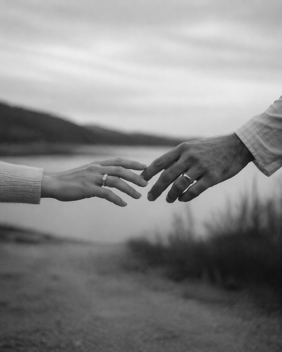 Couple reaching for each other's hands wearing a bespoke diamond engagement ring and men's wedding band. Romantic proposal and bridal jewellery photography celebrating love, commitment and handcrafted fine jewellery. Buy today.
