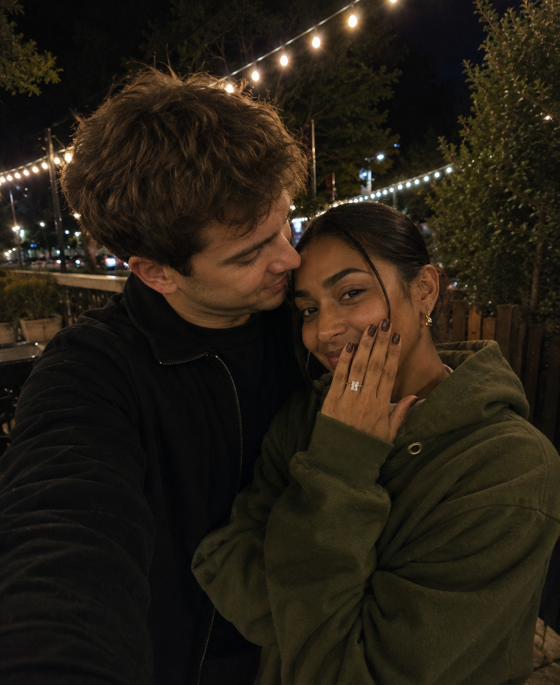 Couple taking a celebratory selfie after a proposal, showcasing a bespoke Radiant diamond engagement ring beneath evening lights. Romantic engagement photography capturing love, commitment and modern proposals.