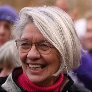Woman with short gray hair and glasses smiling outdoors in a crowd.