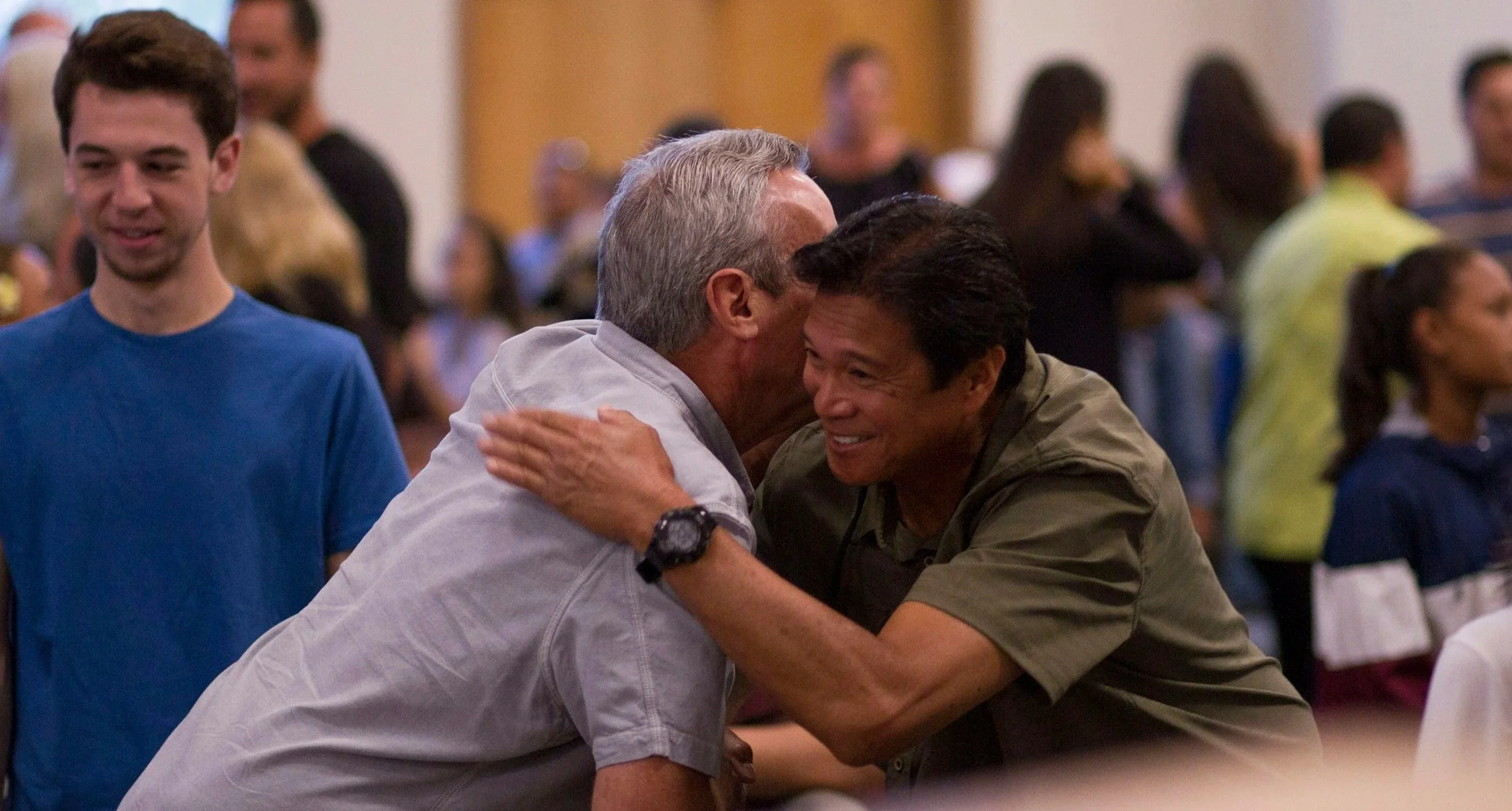 Two men are hugging and smiling at each other in a crowded indoor setting, with several other people visible in the background.