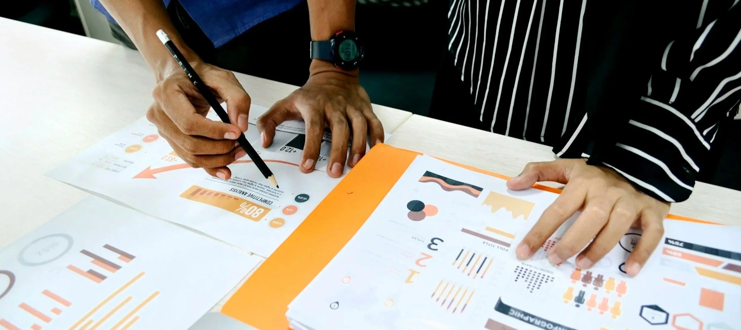 Two people are discussing business charts and infographics on printed sheets, with one person pointing at a document with a pen, on a white table.
