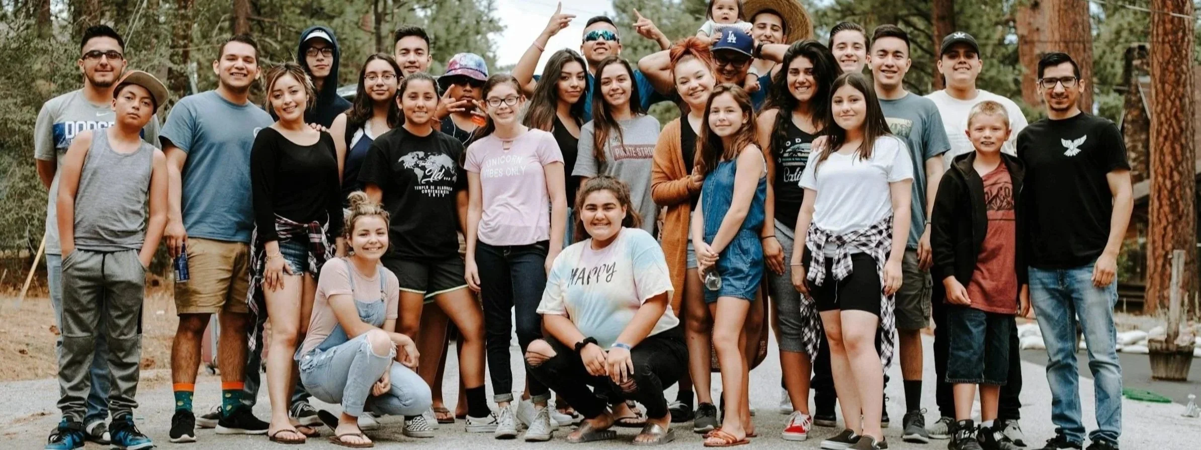 A large group of children and teenagers gathering outdoors in a wooded area, smiling at the camera.