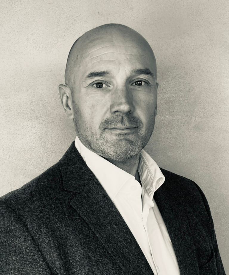 A black and white portrait of a man, wearing a white shirt and a dark blazer, standing against a plain wall.