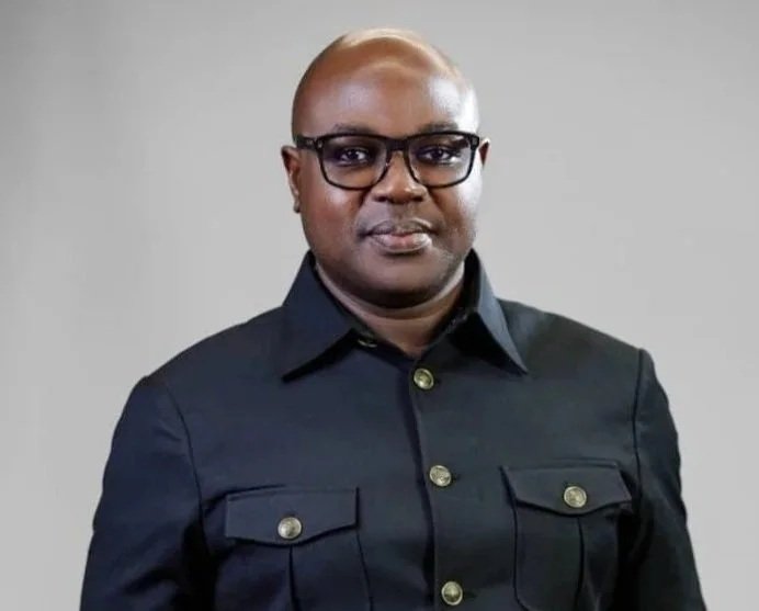 A man wearing glasses, and a black button-up jacket, standing against a plain gray background.