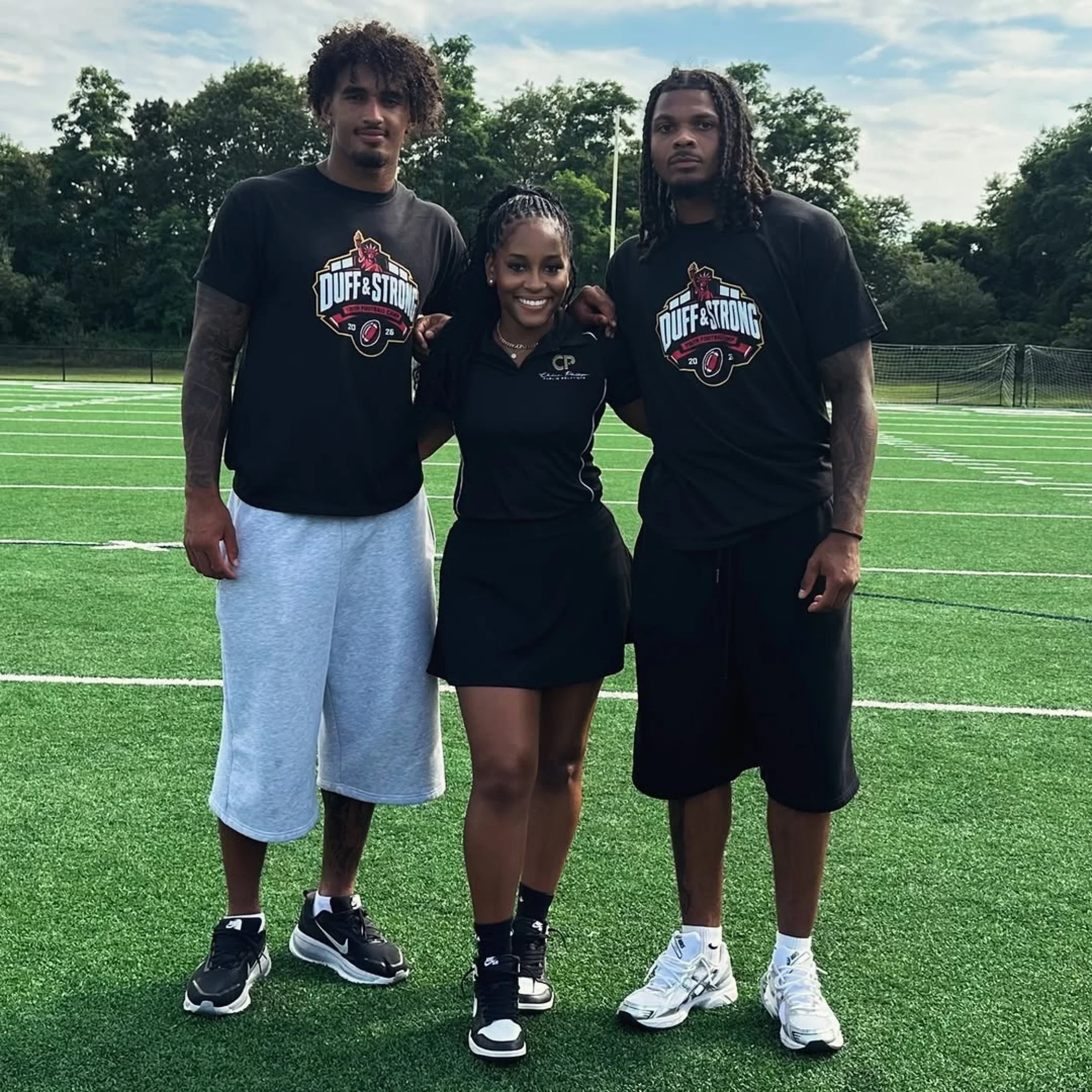Three young adults standing on a football field, wearing black shirts with a 'Duff & Strong' logo, smiling and posing for a photo with their arms around each other, trees and a cloudy sky in the background.