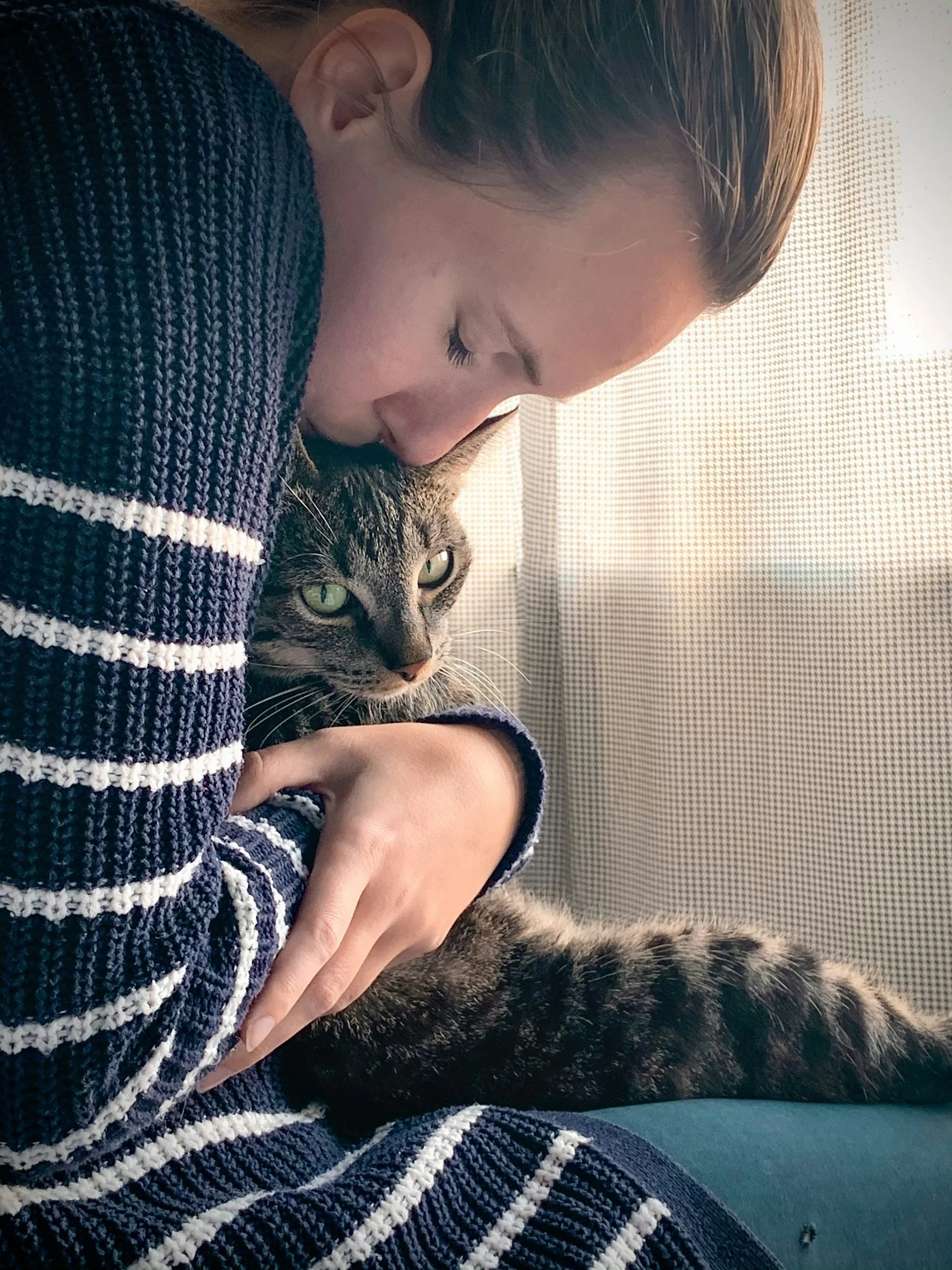 A person with closed eyes wearing a striped sweater, holding a tabby cat with green eyes and a striped coat, both resting peacefully.