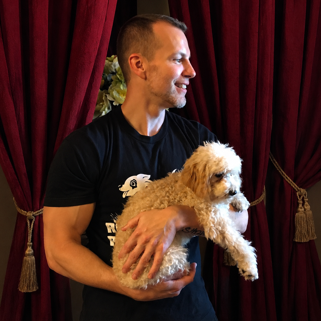 A man with short hair and a beard holding a small, curly-haired puppy in front of red curtains.