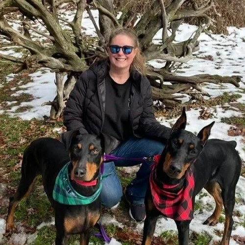 A woman smiling in sunglasses kneeling outdoors with two Doberman dogs, one wearing a green plaid bandana and the other a red plaid bandana, in a snowy landscape with a large bare tree behind them.