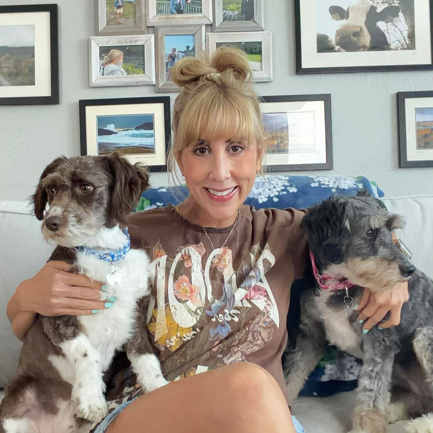 A woman with blonde hair styled in a bun, smiling, holding two small dogs, one brown and white with a blue bandana, and the other gray with a pink collar, inside a room decorated with framed pictures on the wall.
