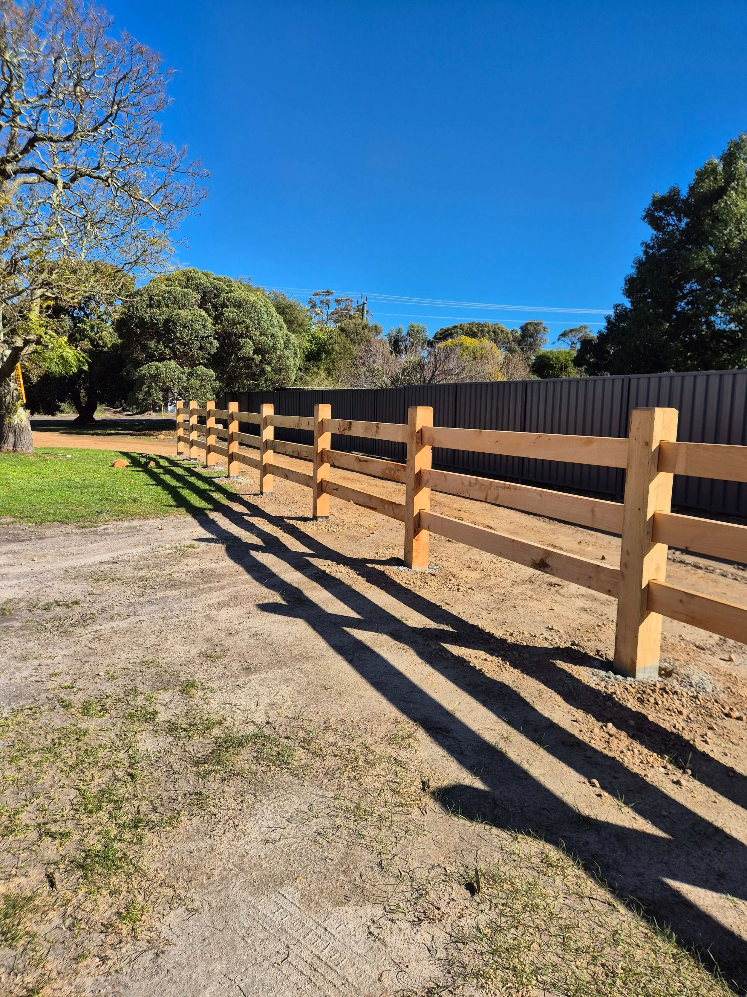 A dirt pathway next to a tidelock post/rail wooden fence and a black metal fence, with trees and a clear blue sky in the background.