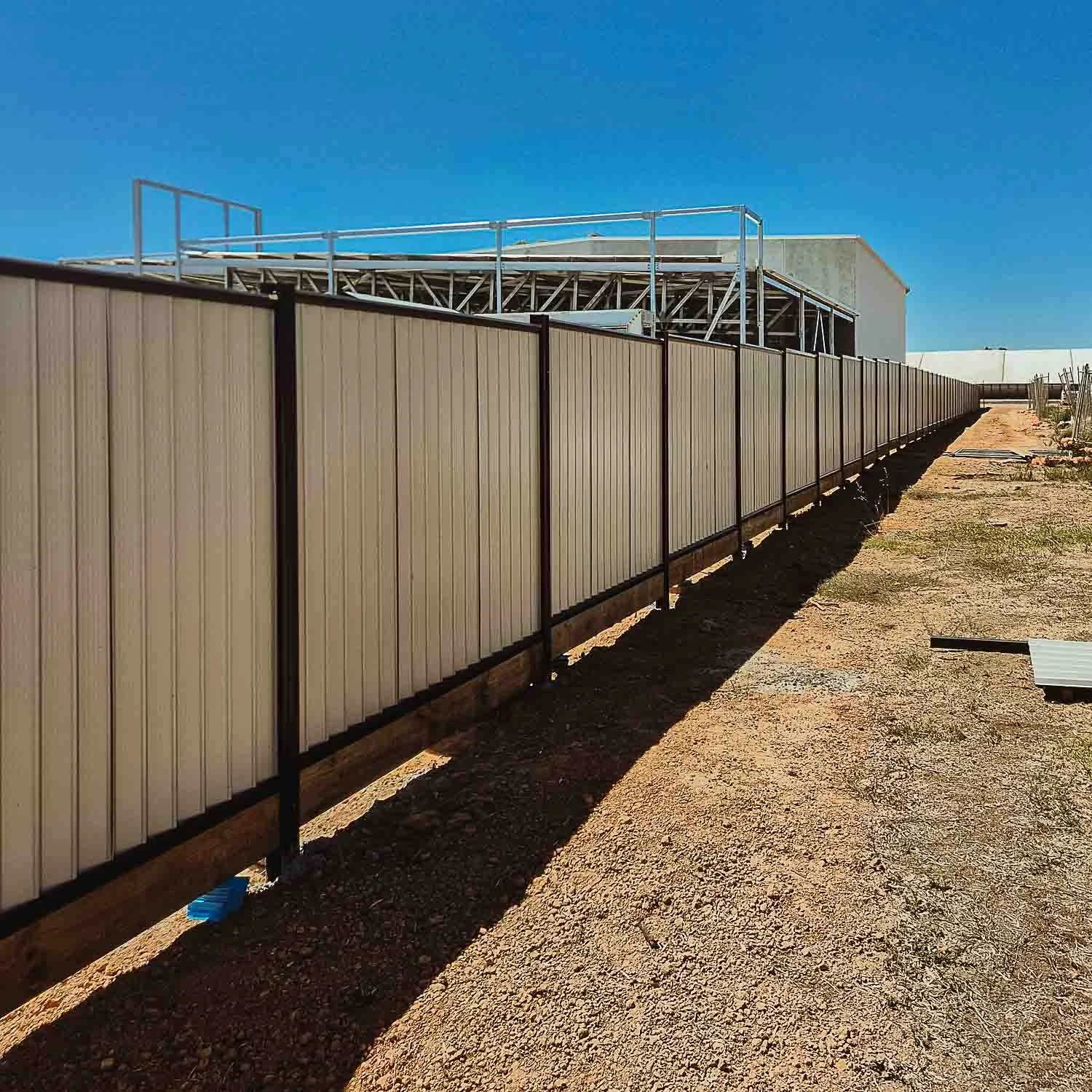 Metal fence running alongside a construction site with a building under construction behind the fence, under a clear blue sky.
