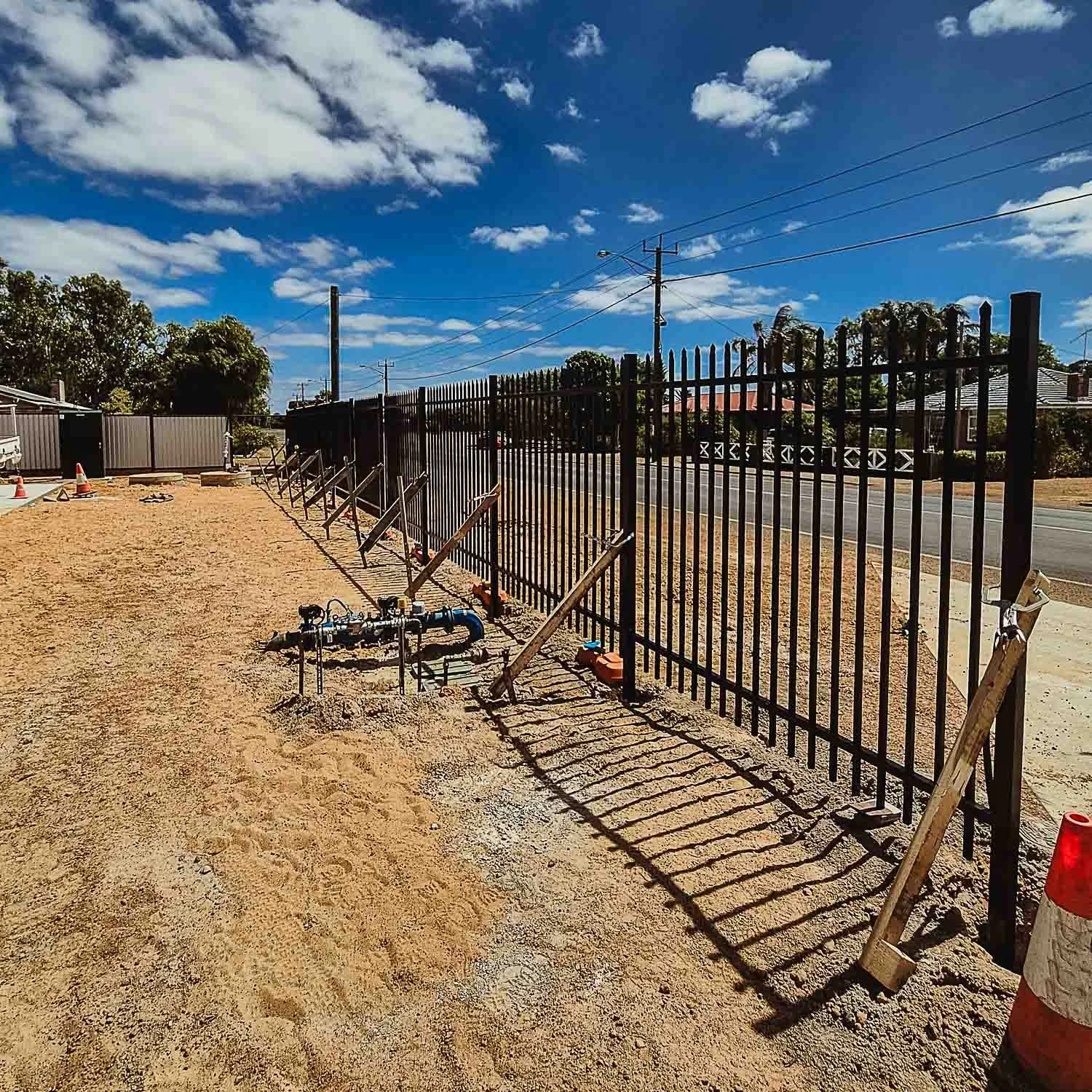 A construction site with a black metal fence, some pipes and construction equipment on sandy ground, residential houses and trees in the background, under a partly cloudy blue sky.