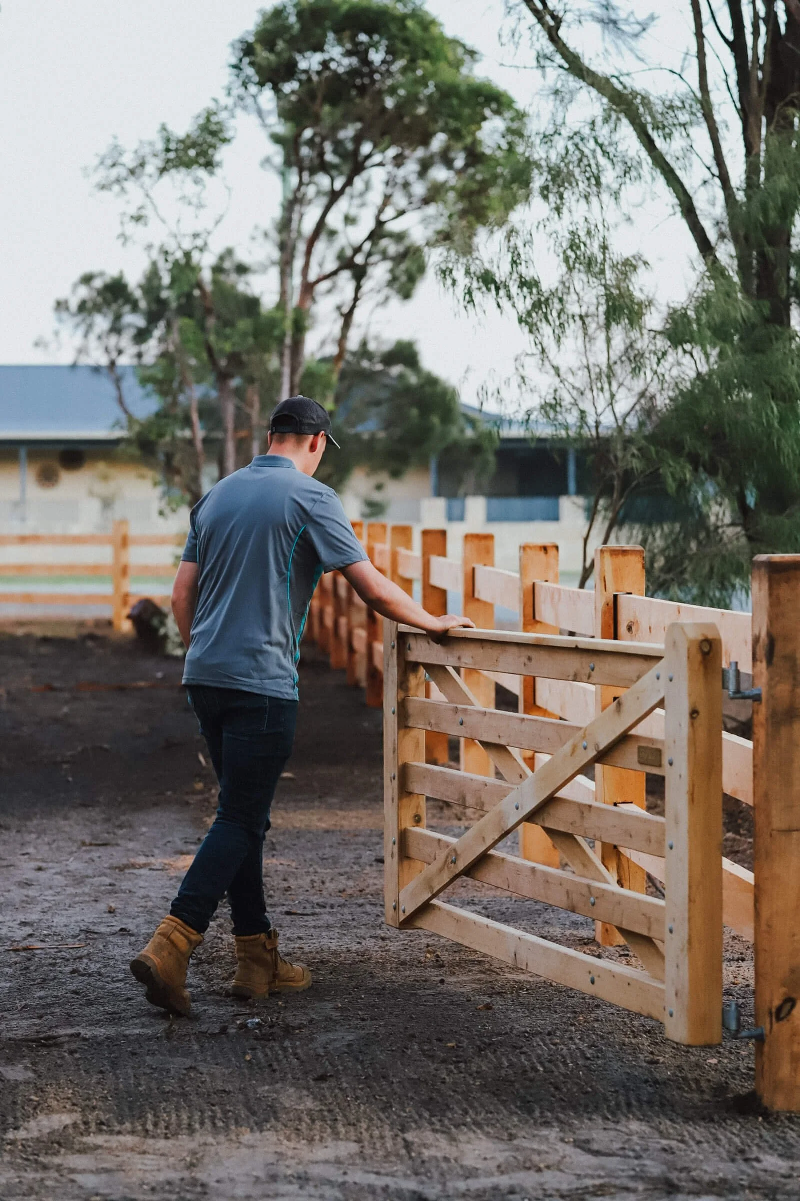 A man in a blue shirt, dark jeans, tan work boots, and a black cap installs a wooden fence on a dirt ground, with trees and a building in the background during daytime.