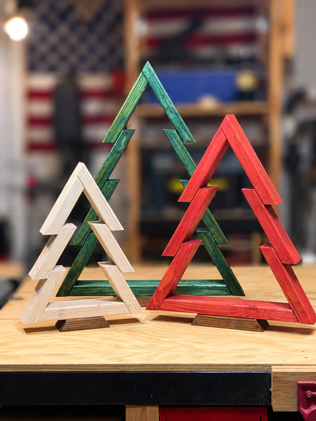 Three wooden Christmas tree decorations painted in white, green, and red, placed on a wooden table in a workshop setting.