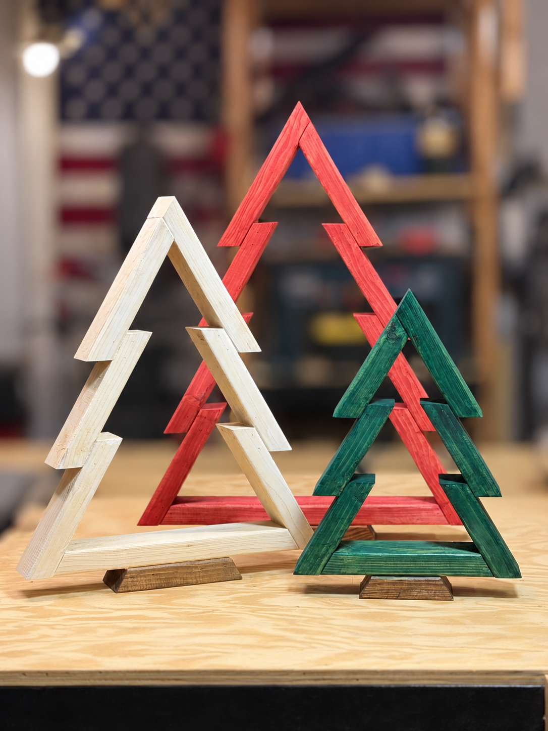 Three wooden Christmas tree decorations painted in red, green, and natural wood color, arranged on a wooden table with a workshop background.
