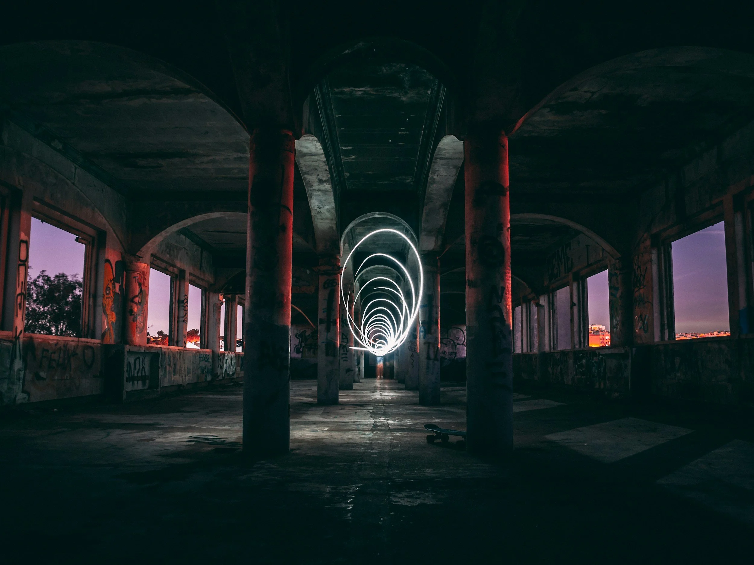 Underpass with graffiti-covered walls, red support columns, and spiral light trails created by long exposure photography, with a skateboard on the ground in front.