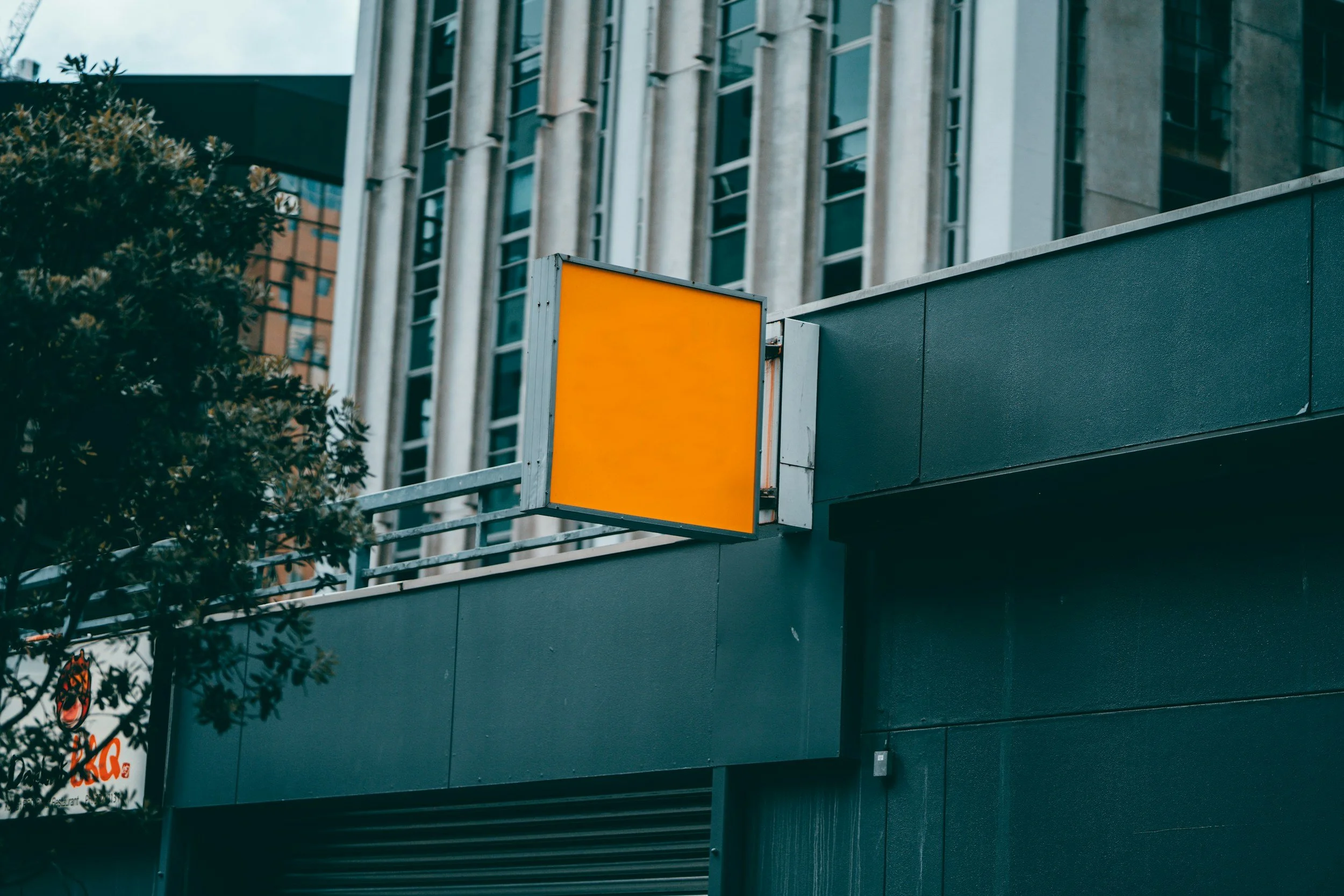An urban building exterior with modern architecture, featuring large glass windows and a yellow blank billboard or sign on a gray facade, with a tree partially visible in the foreground.