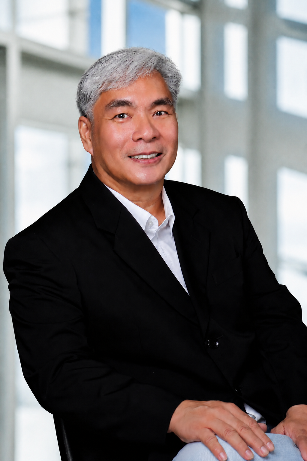 A middle-aged Asian man with gray hair, wearing a black suit and white shirt, sitting with his arms crossed in front of a modern office background with large windows.
