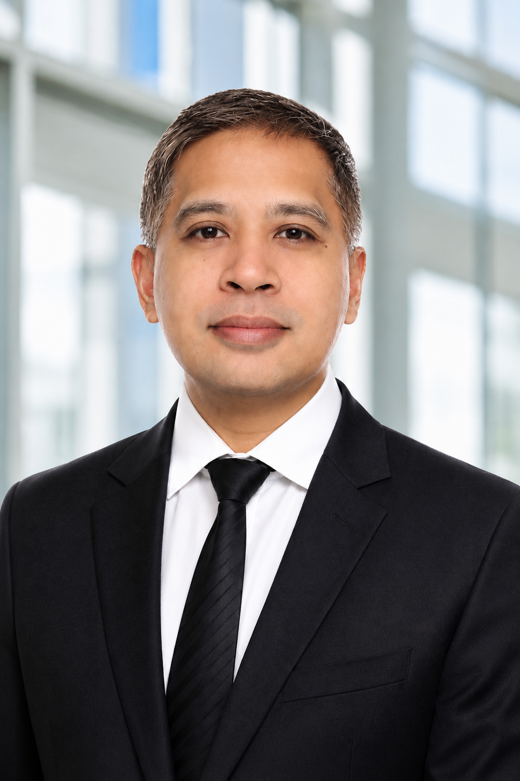 A professional man in a black suit and tie posing indoors with a modern office background.