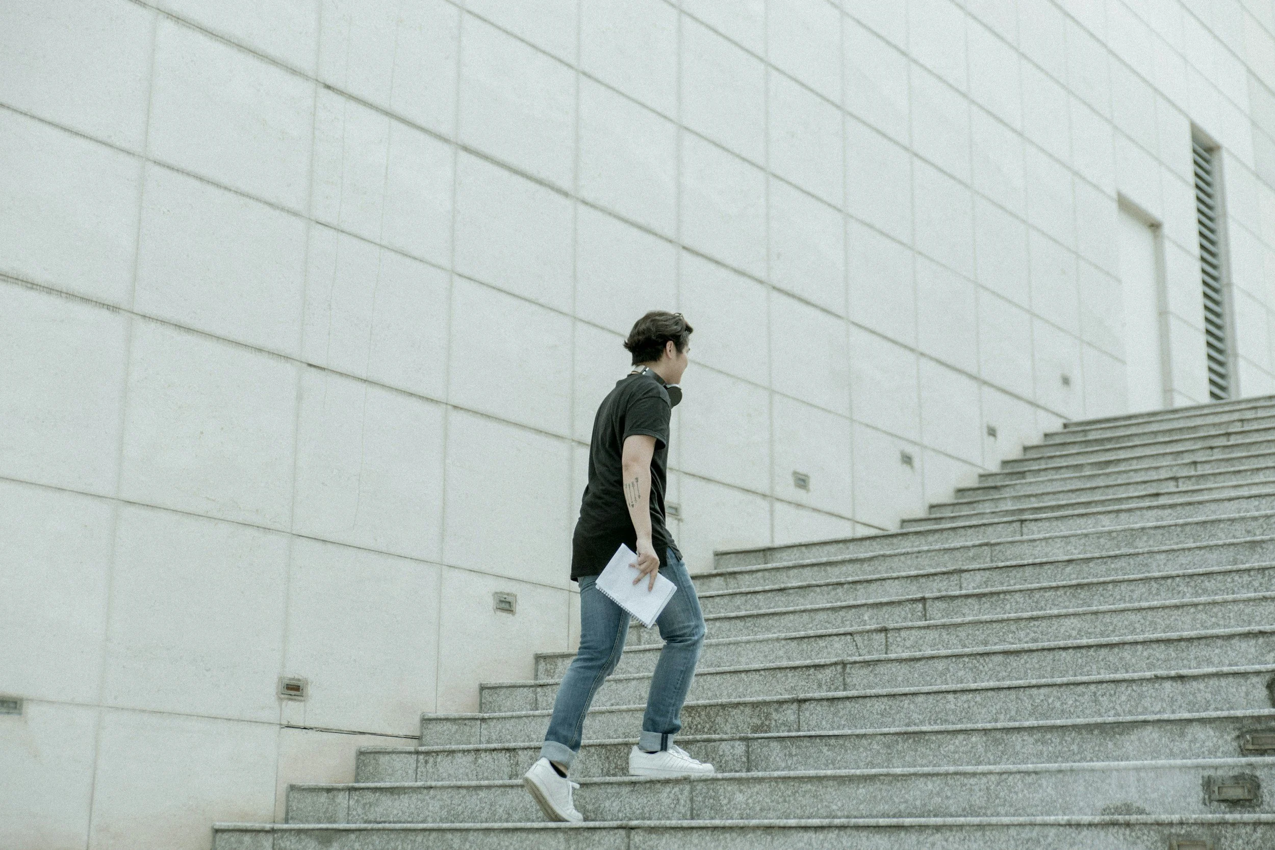 A young man with dark hair wearing a black t-shirt, jeans, and white sneakers walking up concrete stairs holding papers, in front of a large, plain concrete wall with small, rectangular vents.