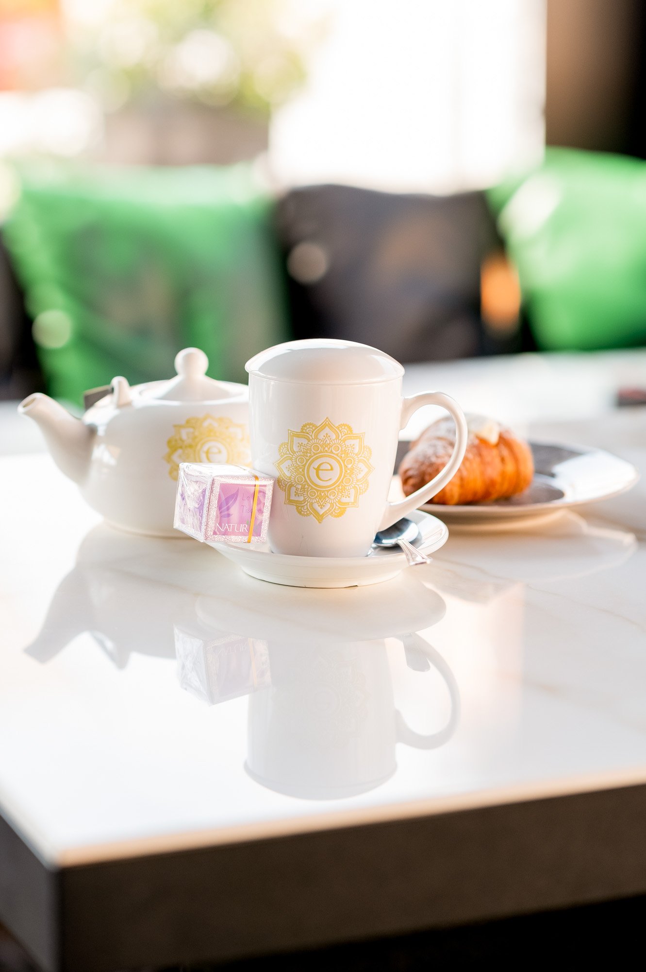 Tazza bianca con decorazione dorata, teiera bianca, pacchetto di tè e dolci su un vassoio su un tavolo con sfondo sfocato di sedie verdi.