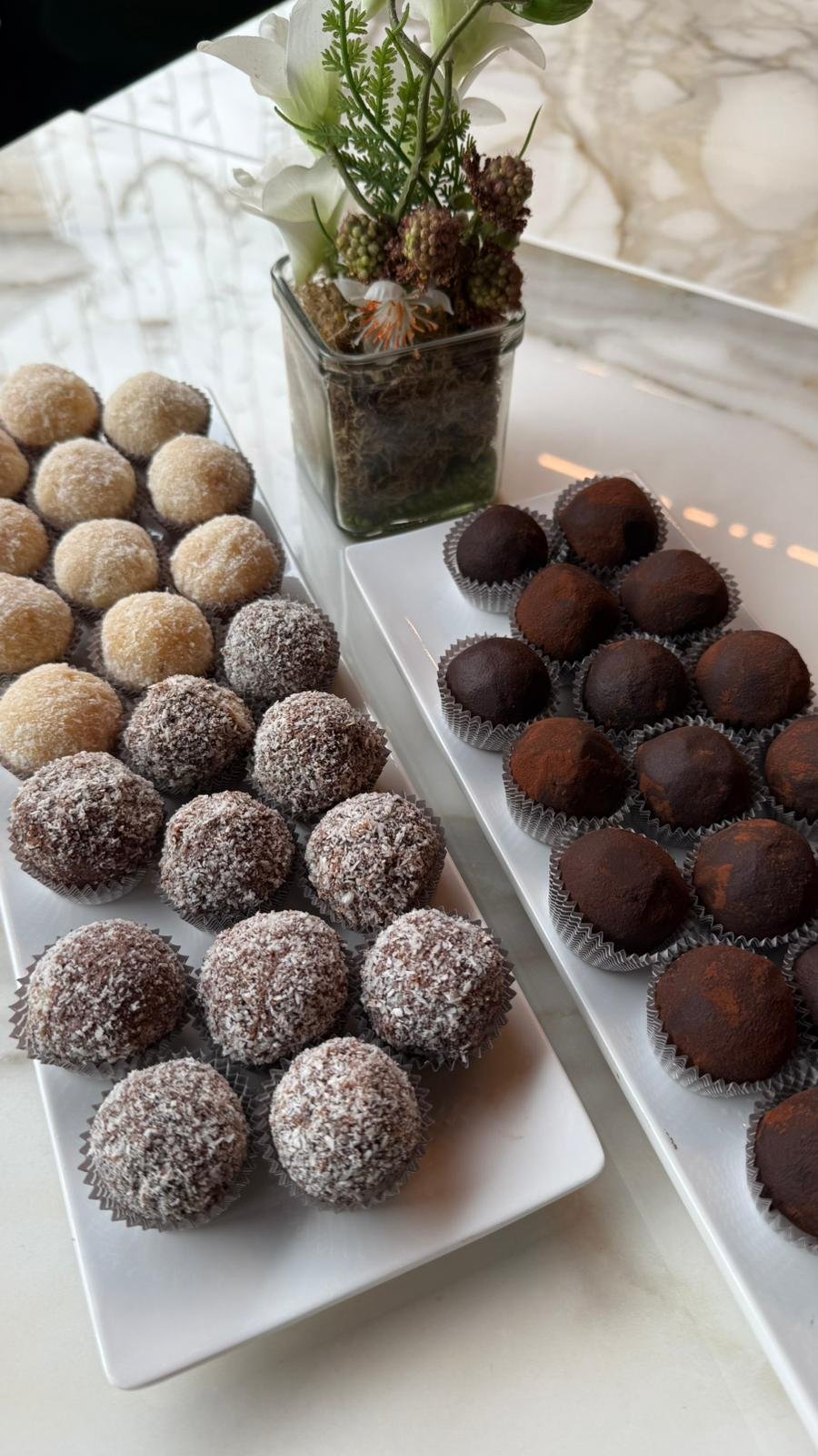Piattini con palline di cioccolato, alcune ricoperte di cocco e altre di cacao, accanto a un fiore bianco in vaso.