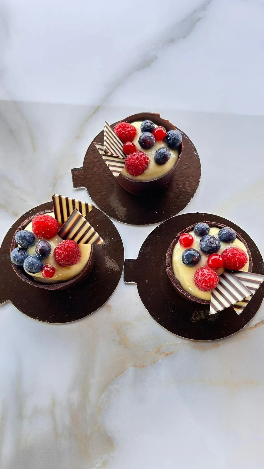 Tre dolci al cioccolato decorati con frutti di bosco freschi e cioccolato bianco e fondente, presentati su una superficie di marmo.