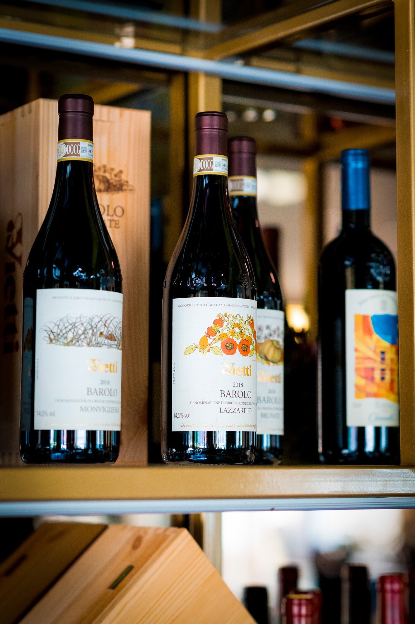 Bottiglie di vino rosso sulla mensola di una vineria, con diverse etichette e un packaging in legno sullo sfondo.