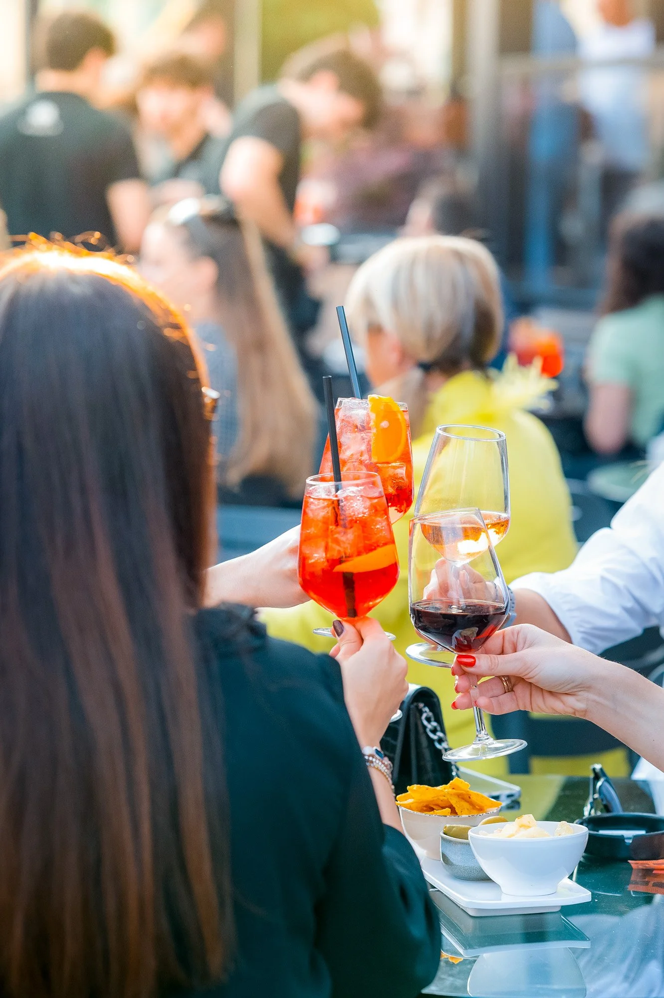 Persone che brindano con bicchieri di vino e bevande colorate durante un evento sociale, con cibo sul tavolo.