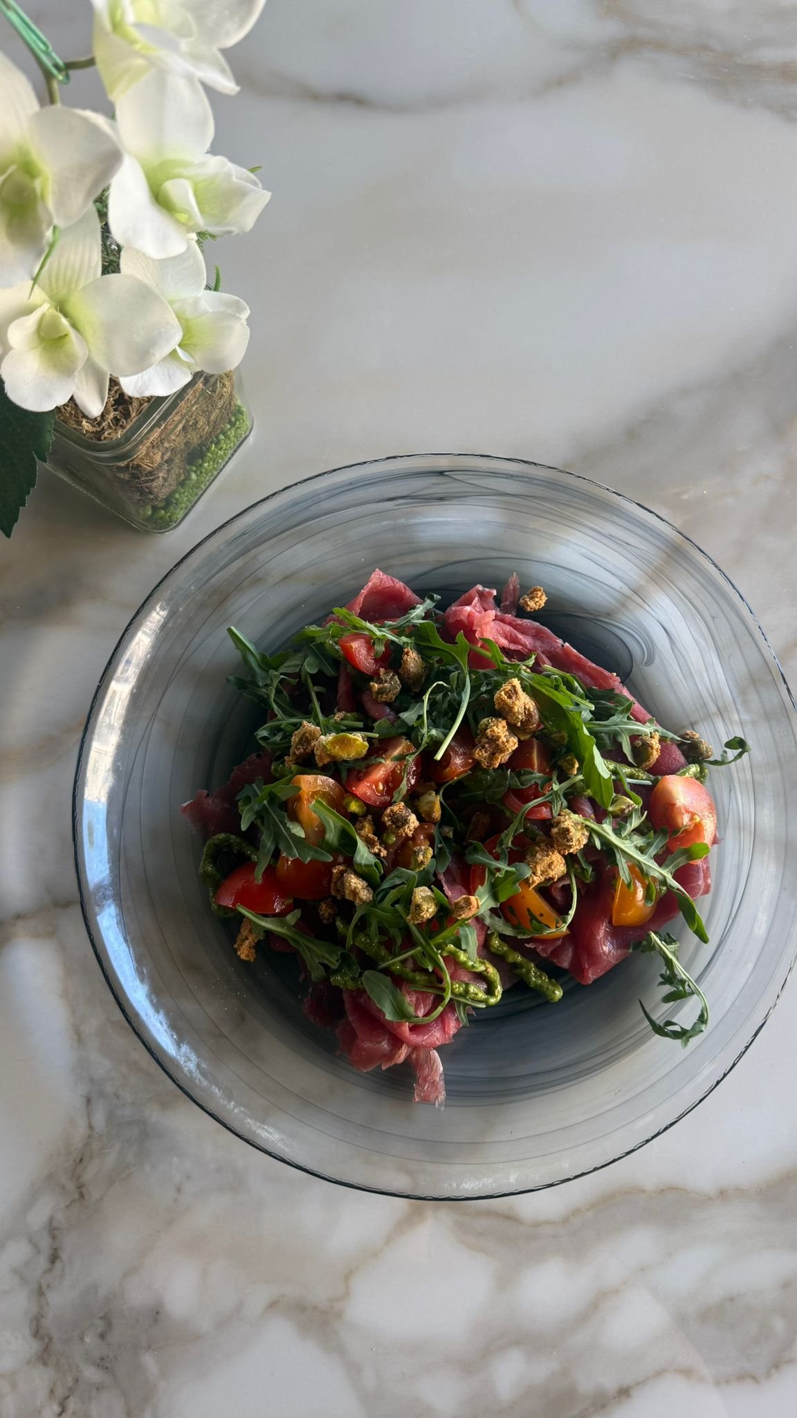 Insalata con pomodorini, rucola e speck in una ciotola di vetro, a lato un vaso con fiori bianchi.