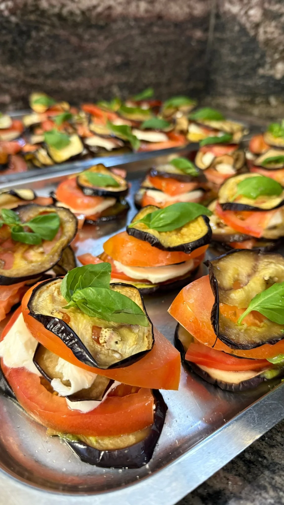 Verdure grigliate con pomodori, melanzane, formaggio e basilico su un vassoio di metallo.