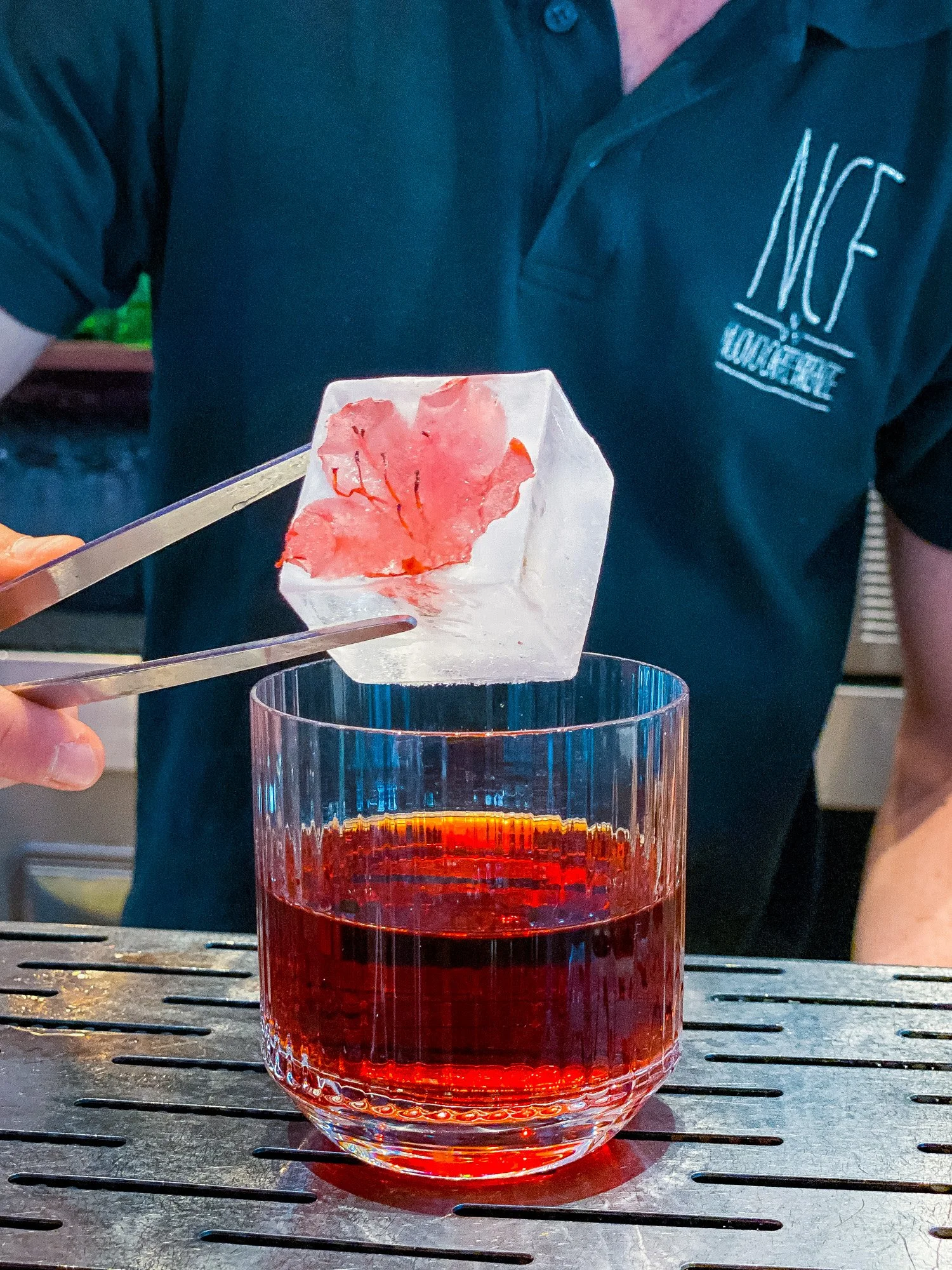 Un bicchiere di vetro contiene un liquido rosso acceso. Sopra la superficie del liquido c'è una sostanza di colore rosa chiaro, forse un pezzo di carne o una pietanza. Una mano con pinze sta sollevando la sostanza dal bicchiere.