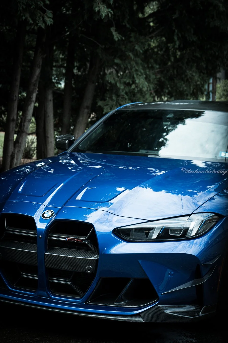 Front view of a blue BMW M8 car parked outdoors with trees in the background.