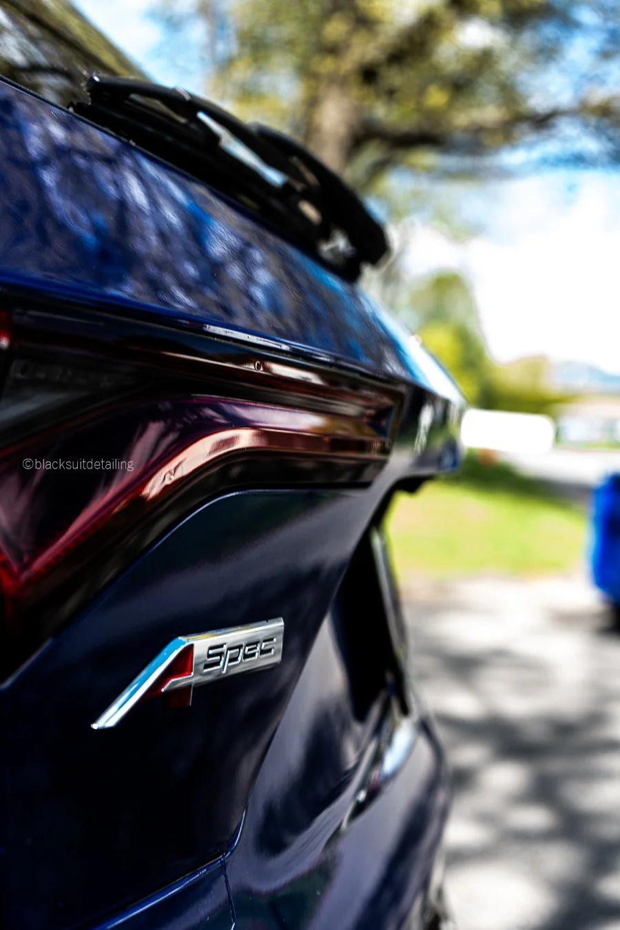 Close-up of a blue car's rear with an 'A-Spec' badge, blurred background with trees and a driveway.