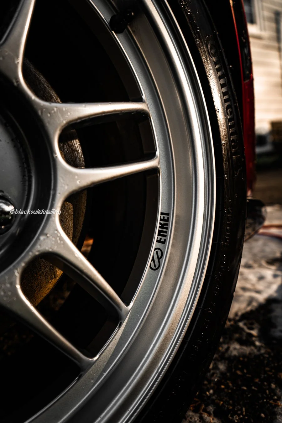 Close-up of a silver Enkei alloy wheel with a black tire, water droplets on the surface, and a partial view of a red car body.