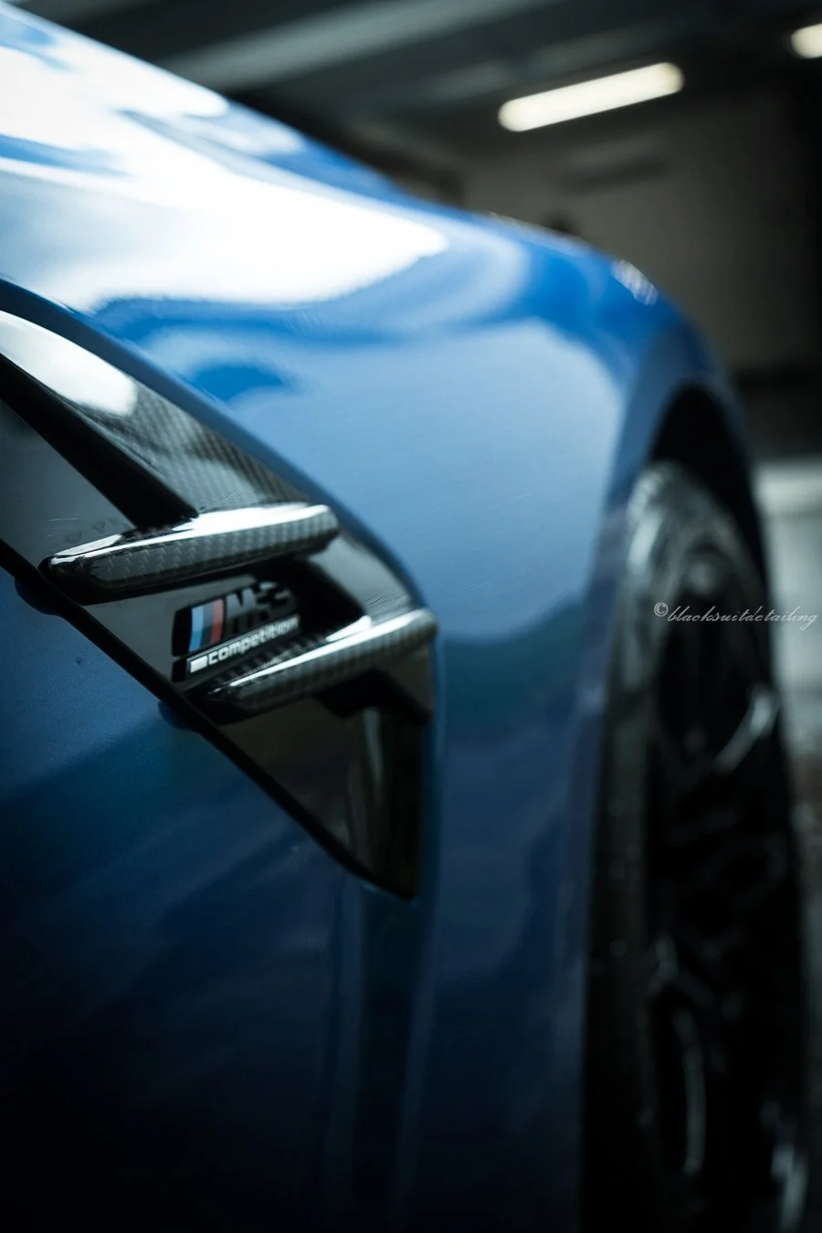 Close-up of a blue sports car's side vent with carbon fiber accents and BMW M logo, inside a garage.