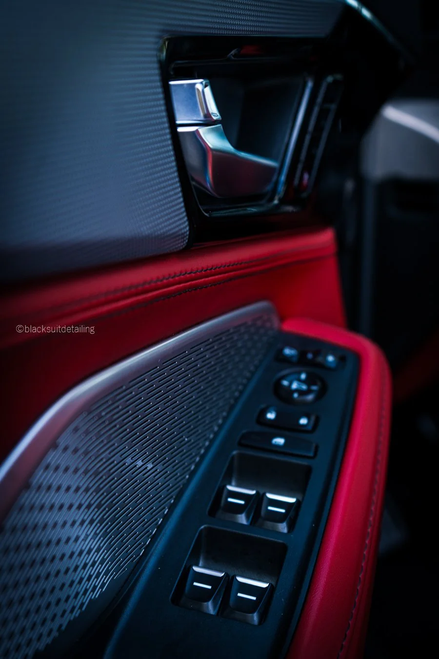 Close-up of a car door interior featuring a metallic speaker grille, window control buttons, and red leather stitching.