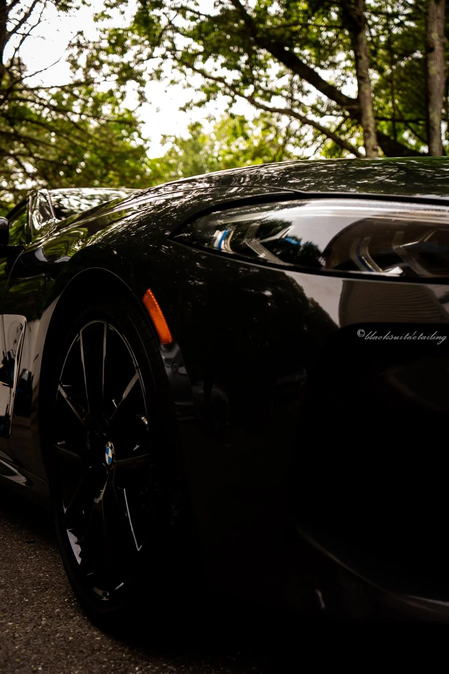 Close-up of the front part of a shiny black sports car with a reflection of trees and the sky on its surface, parked outdoors.