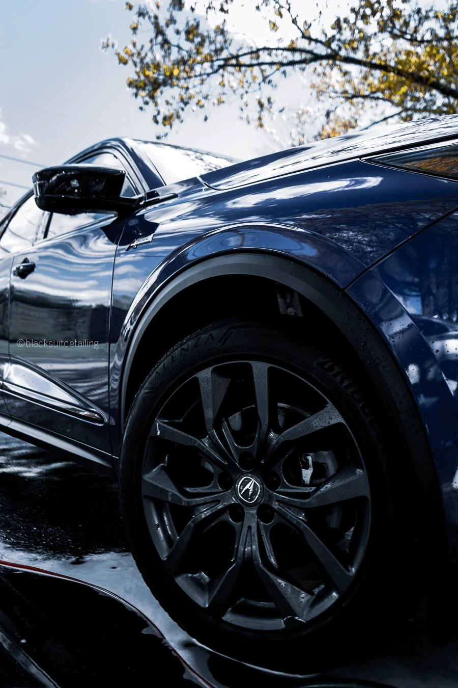 Close-up of a blue car's front side, including the wheel and tire, with reflections on its surface. The background shows a tree with yellow leaves and a cloudy sky.