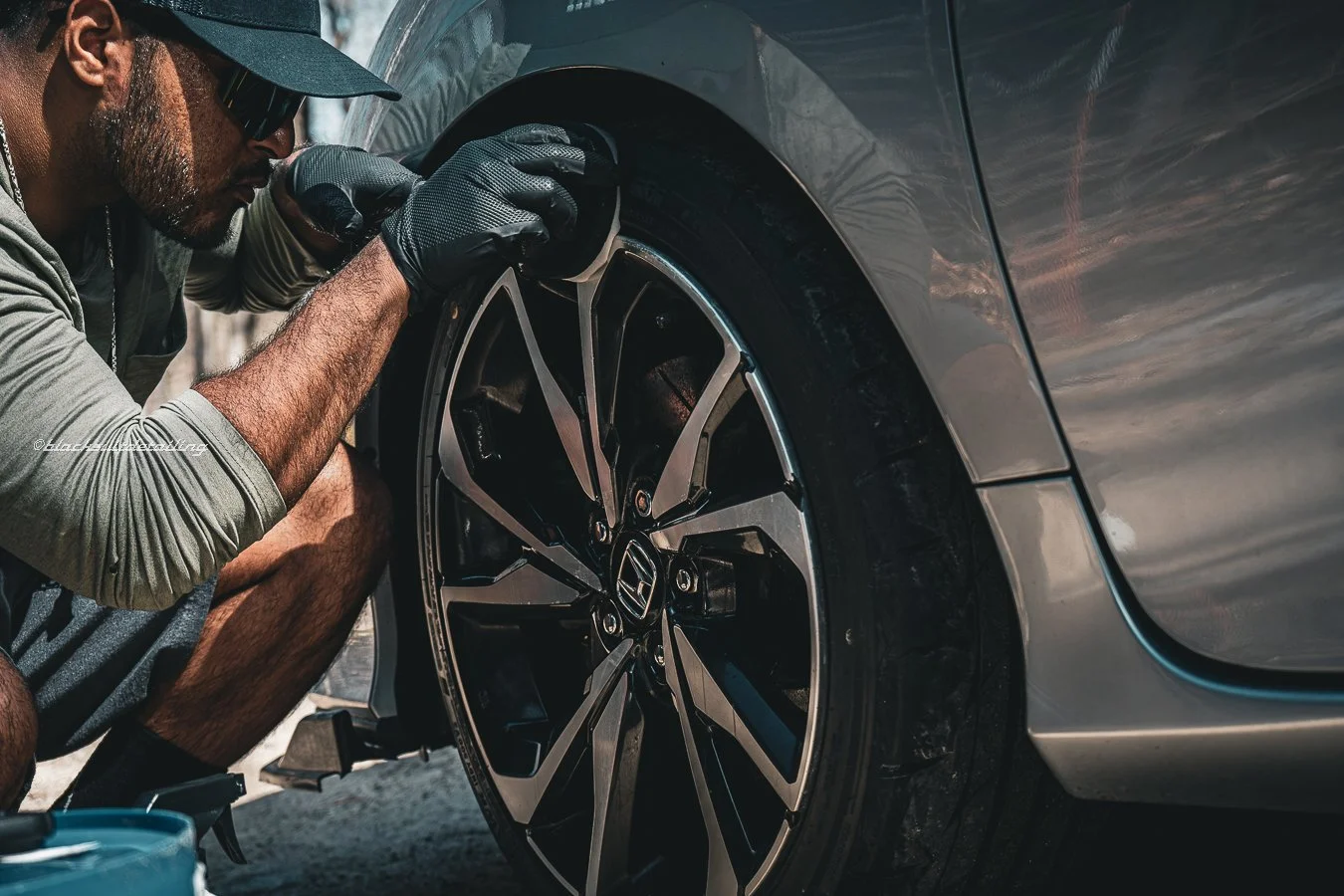 A man wearing a cap, sunglasses, a gray long-sleeve shirt, and black gloves is fixing or inspecting the wheel of a gray car, possibly a Honda, outdoors on a sunny day.