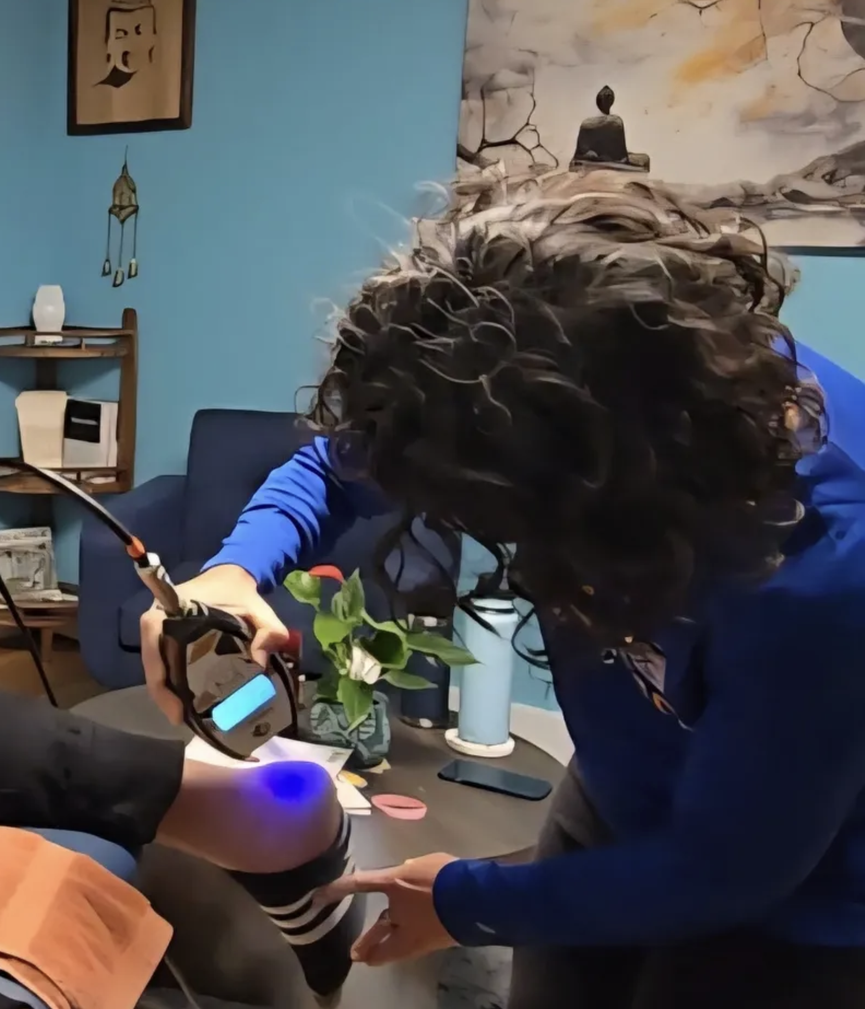 Person with curly hair working on a project with a soldering iron at a desk in a room with blue walls, a plant, and artwork on the wall.