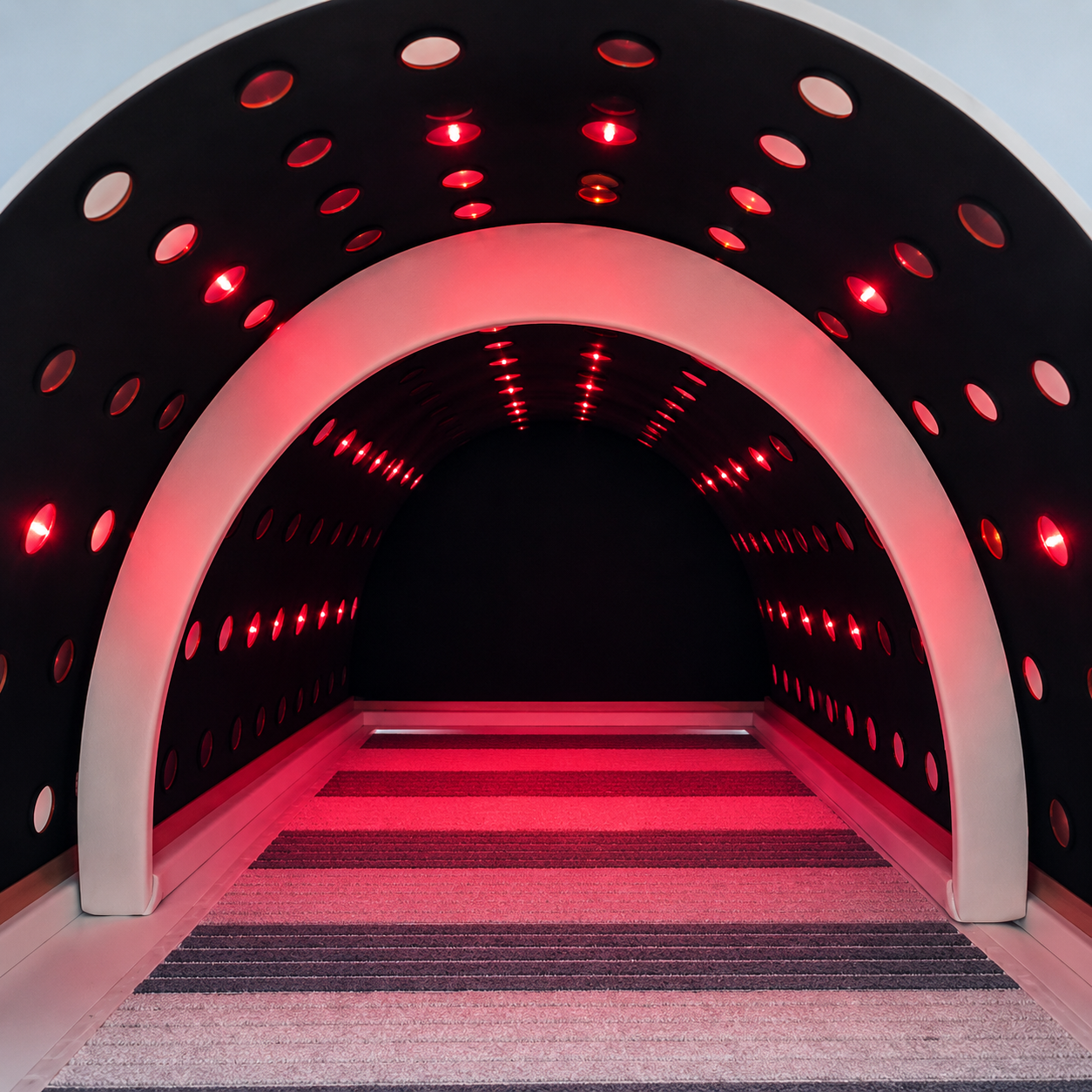 A futuristic tunnel with curved black walls featuring evenly spaced circular holes and red LED lights, with a striped carpeted floor leading into darkness.
